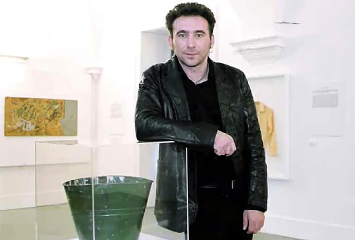 Guy Tarrant dressed in black, leaning against one of his artworks in a gallery